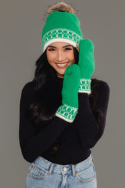 A woman smiles at the camera, wearing the Green & Cream Patterned Knit Pom Hat by Panache Apparel Collection and matching green mittens, both featuring white patterns. She is dressed in a black top and light blue jeans against a plain gray background.