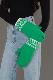 A person in light blue jeans and a black long-sleeve top crosses their arms, showcasing the Panache Apparel Collection Green & Cream Patterned Knit Mittens against a gray background.