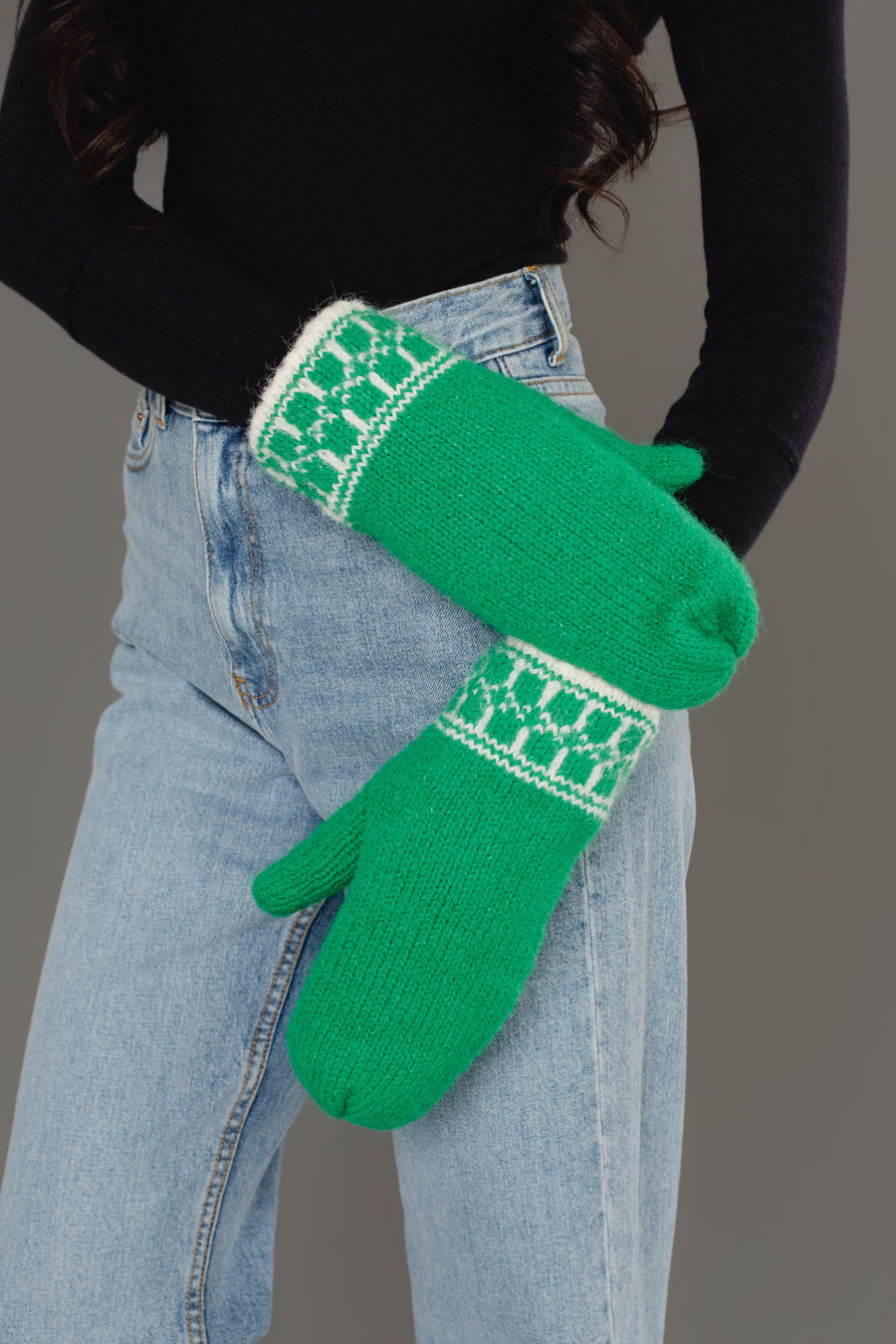 A person in light blue jeans and a black long-sleeve top crosses their arms, showcasing the Panache Apparel Collection Green & Cream Patterned Knit Mittens against a gray background.