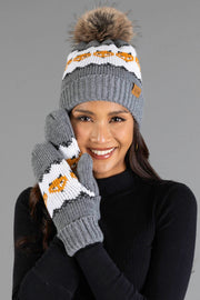 A smiling woman wears the Panache Apparel Collection’s Gray with Orange Fox Pattern Knit Pom Hat, paired with matching mittens, while posing against a plain gray background.