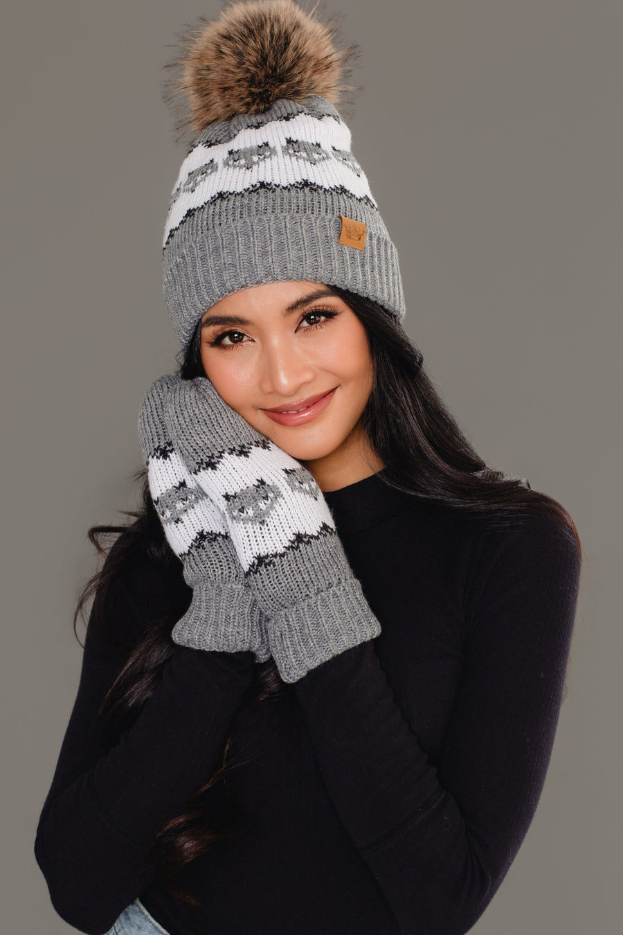 A smiling woman models the Panache Apparel Collection’s Gray with Gray Fox Pattern Knit Pom Hat, posing with one hand on her cheek against a plain grey background.