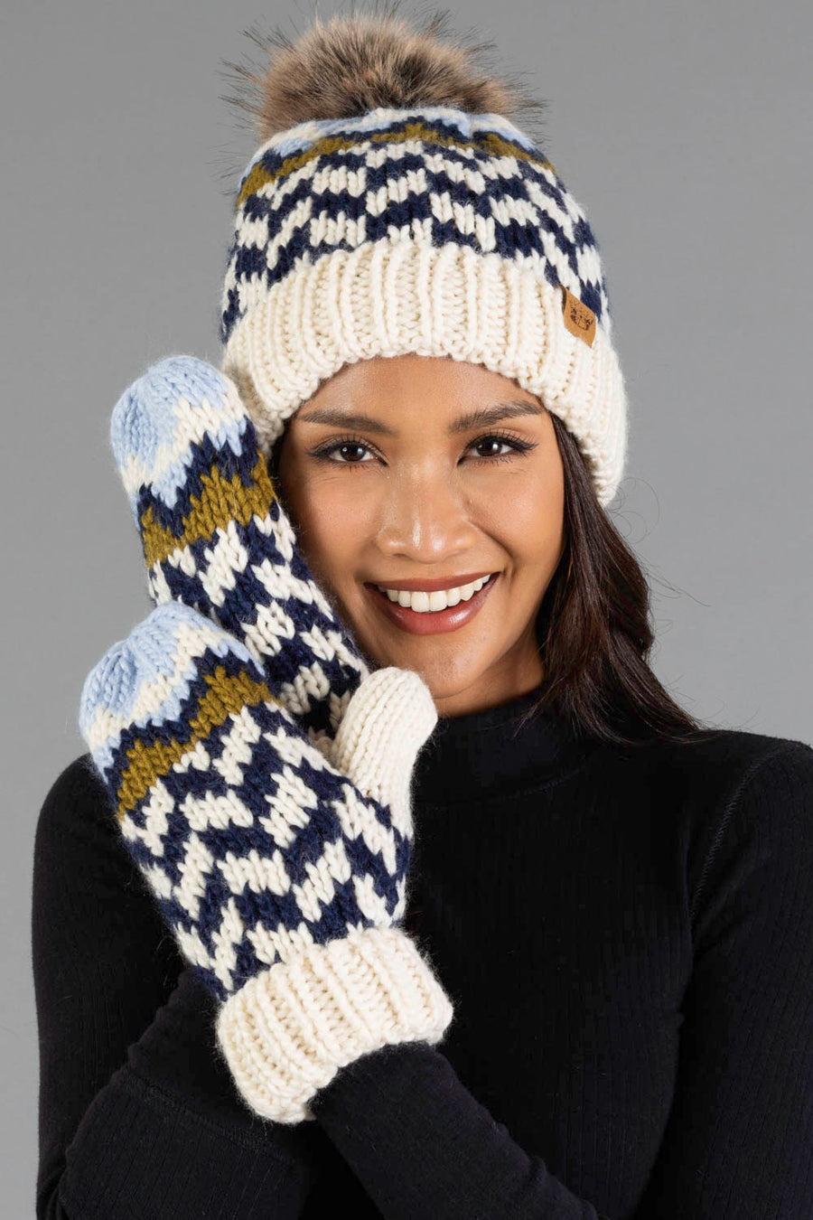 A woman smiles while wearing the Panache Apparel Collection Beige, Navy & Light Blue Patterned Pom Hat and matching fleece-lined mittens. The cozy knit hat, finished with a faux fur pom-pom, and coordinating cream, blue, green, and navy mittens are showcased as she holds her hands near her face against a gray background.