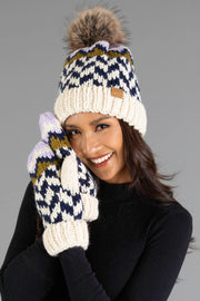 A woman with long brown hair, dressed in a black top, smiles as she holds her gloved hands near her face, wearing the Panache Apparel Collection Beige, Navy & Lilac Patterned Pom Hat with matching mittens.