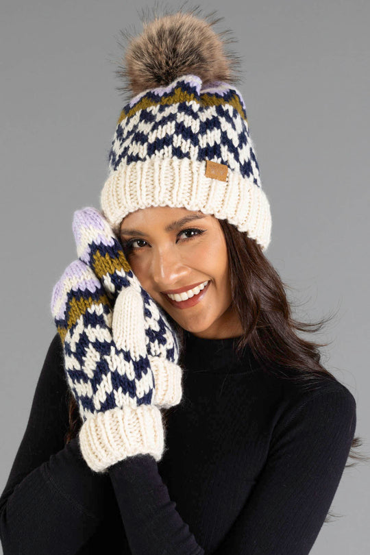 A woman with long brown hair, dressed in a black top, smiles as she holds her gloved hands near her face, wearing the Panache Apparel Collection Beige, Navy & Lilac Patterned Pom Hat with matching mittens.