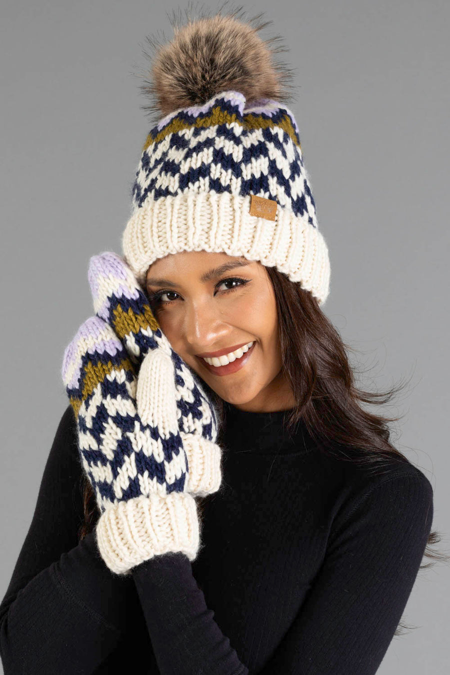 A woman with long brown hair, dressed in a black top, smiles as she holds her gloved hands near her face, wearing the Panache Apparel Collection Beige, Navy & Lilac Patterned Pom Hat with matching mittens.