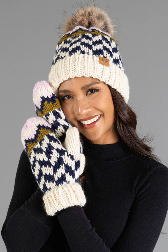 Smiling woman wearing the Panache Apparel Collection Beige, Navy & Light Pink Patterned Pom Hat and matching knit mittens, dressed in a black top and posing in front of a gray background.