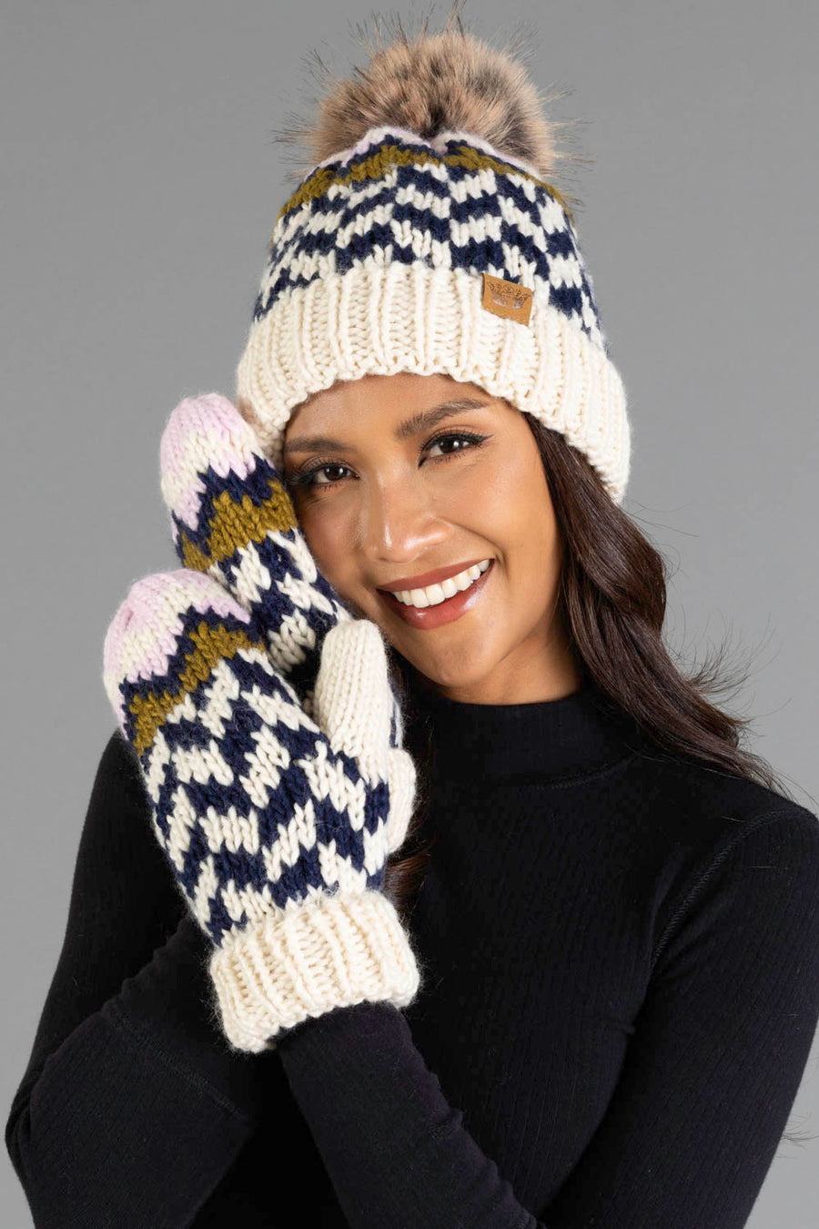 Smiling woman wearing the Panache Apparel Collection Beige, Navy & Light Pink Patterned Pom Hat and matching knit mittens, dressed in a black top and posing in front of a gray background.