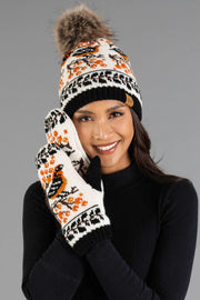 A smiling person wears the Panache Apparel Collection Black & Cream Bird Pattern Knit Pom Hat with a fur pom-pom, paired with matching mittens featuring a bird and orange berry design, set against a gray background.
