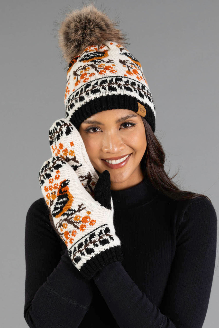 A smiling person wears the Panache Apparel Collection Black & Cream Bird Pattern Knit Pom Hat with a fur pom-pom, paired with matching mittens featuring a bird and orange berry design, set against a gray background.