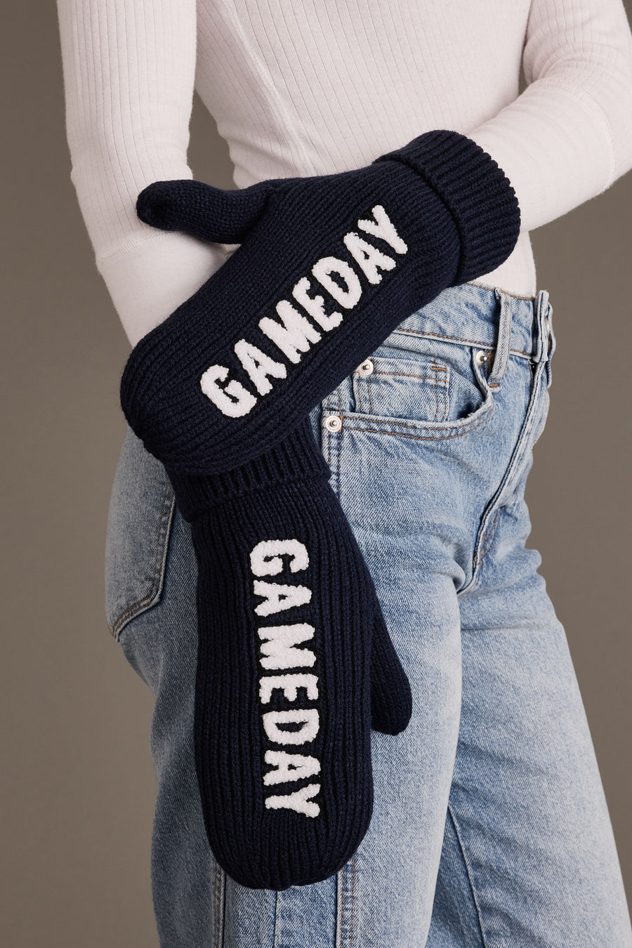 A person wearing a white ribbed top and blue jeans holds the Panache Apparel Collection Navy & White Game Day Mittens, which are fleece-lined and feature “GAMEDAY” written in white letters.