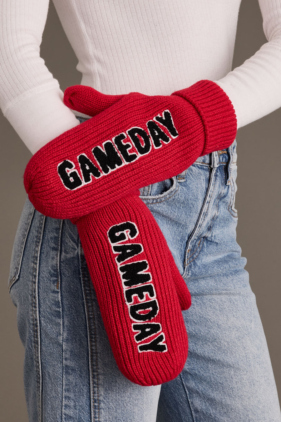 A person wearing Panache Apparel Collection’s Red & Black Game Day Mittens, featuring bold black and white lettering, paired with a white ribbed shirt and blue jeans, stands against a neutral background.