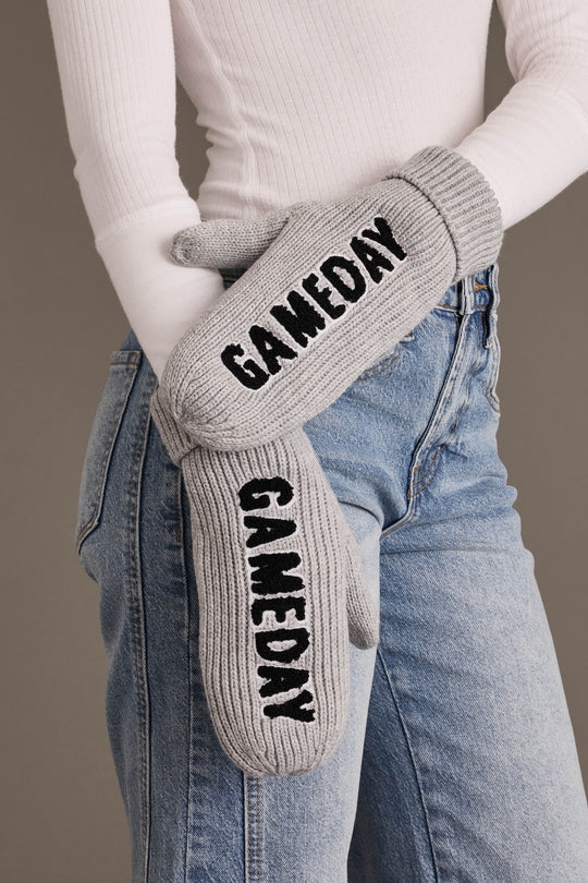 A person wearing light-wash jeans and a white ribbed top holds the Gray & Black Game Day Mittens from Panache Apparel Collection—gray fleece-lined knit mittens featuring “GAMEDAY” in bold black letters on each mitten.