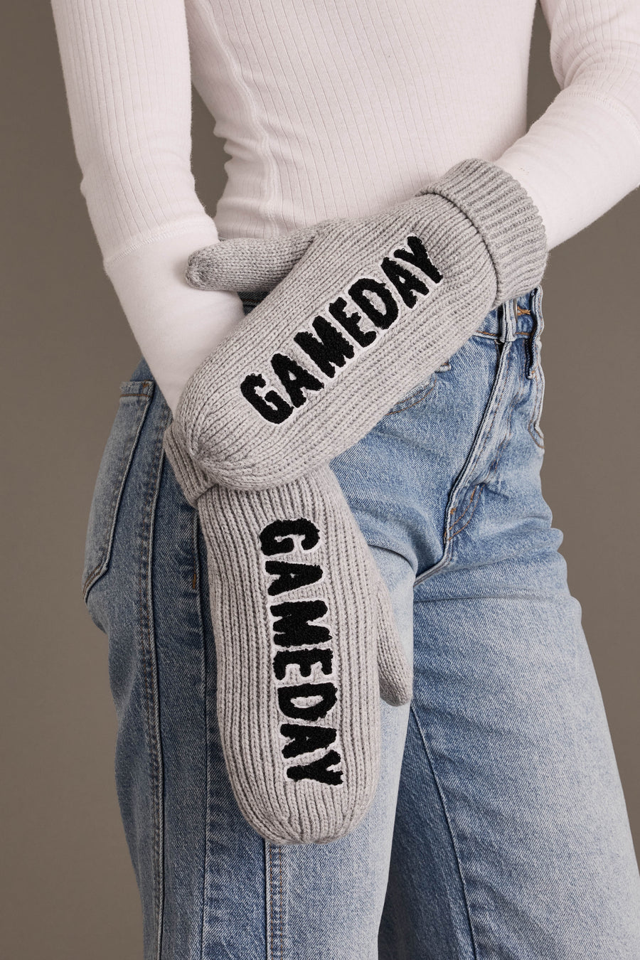 A person wearing light-wash jeans and a white ribbed top holds the Gray & Black Game Day Mittens from Panache Apparel Collection—gray fleece-lined knit mittens featuring “GAMEDAY” in bold black letters on each mitten.