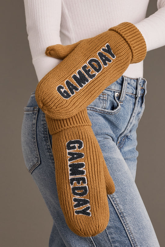 A person wearing Panache Apparel Collection’s Camel & Gray Game Day Mittens, fleece-lined for extra warmth, featuring the word GAMEDAY in bold black and white letters—styled with blue jeans and a white ribbed sweater—for the perfect chilly weather game day accessories.