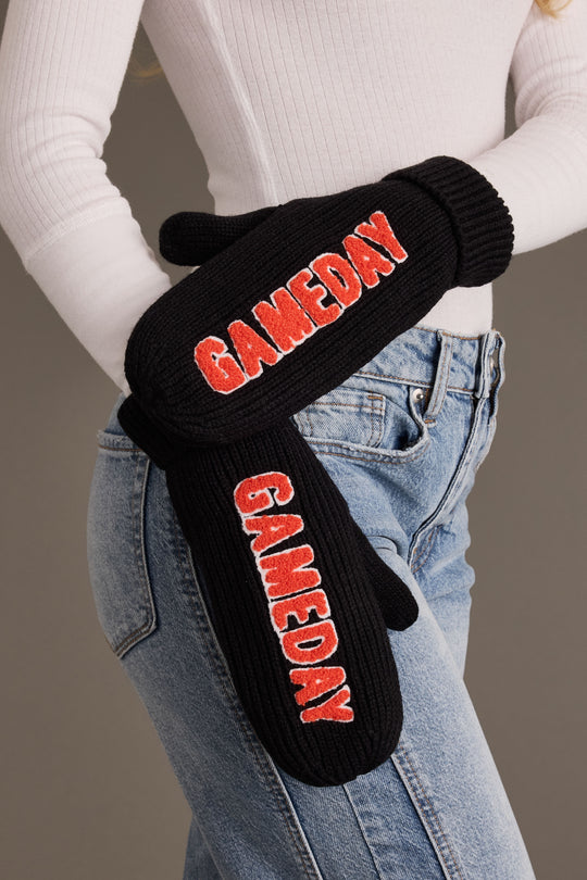 A person wearing a white ribbed sweater and blue jeans with the Panache Apparel Collection Black & Orange Game Day Mittens, featuring the word “GAMEDAY” in red letters on each fleece-lined mitten.