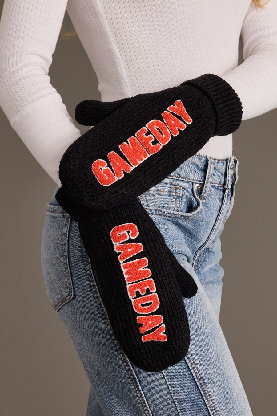 A person wearing a white ribbed sweater and blue jeans with the Panache Apparel Collection Black & Orange Game Day Mittens, featuring the word “GAMEDAY” in red letters on each fleece-lined mitten.