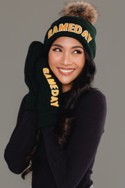 A smiling woman in a black long-sleeve shirt wears the Panache Apparel Collection Green & Gold Game Day Pom Hat, featuring a fleece-lined interior and yellow "GAMEDAY" lettering, along with matching mittens, against a plain gray background.