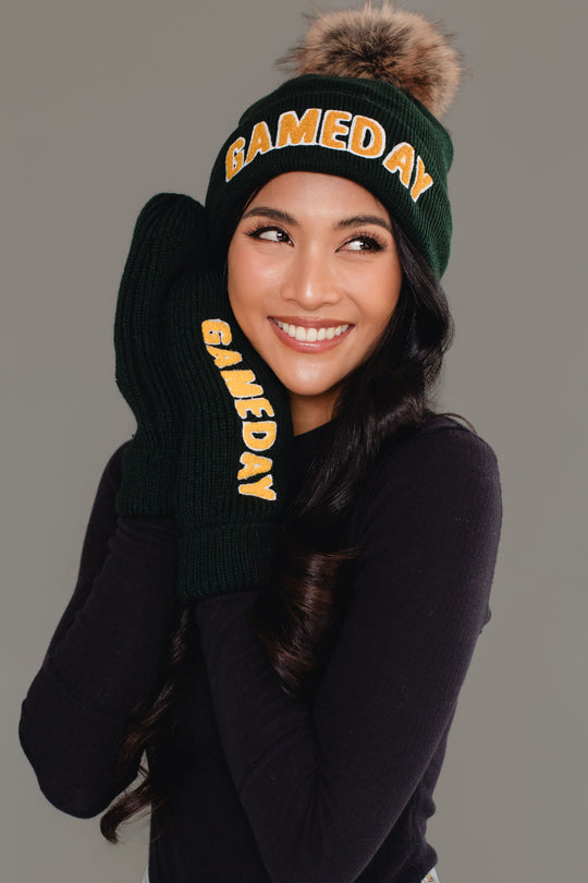 A smiling woman in a black long-sleeve shirt wears the Panache Apparel Collection Green & Gold Game Day Pom Hat, featuring a fleece-lined interior and yellow "GAMEDAY" lettering, along with matching mittens, against a plain gray background.