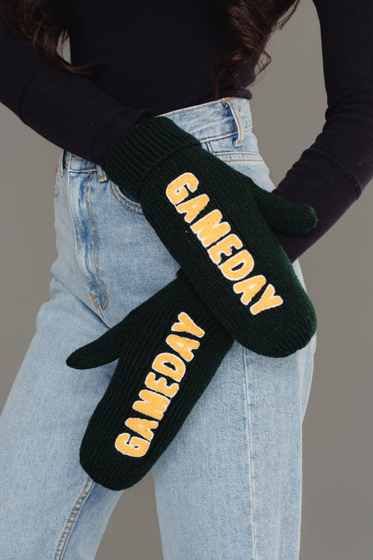 A person wearing a black top and light blue jeans displays the Green & Gold Game Day Mittens by Panache Apparel Collection, featuring the word "GAMEDAY" in yellow and white text on each mitten.