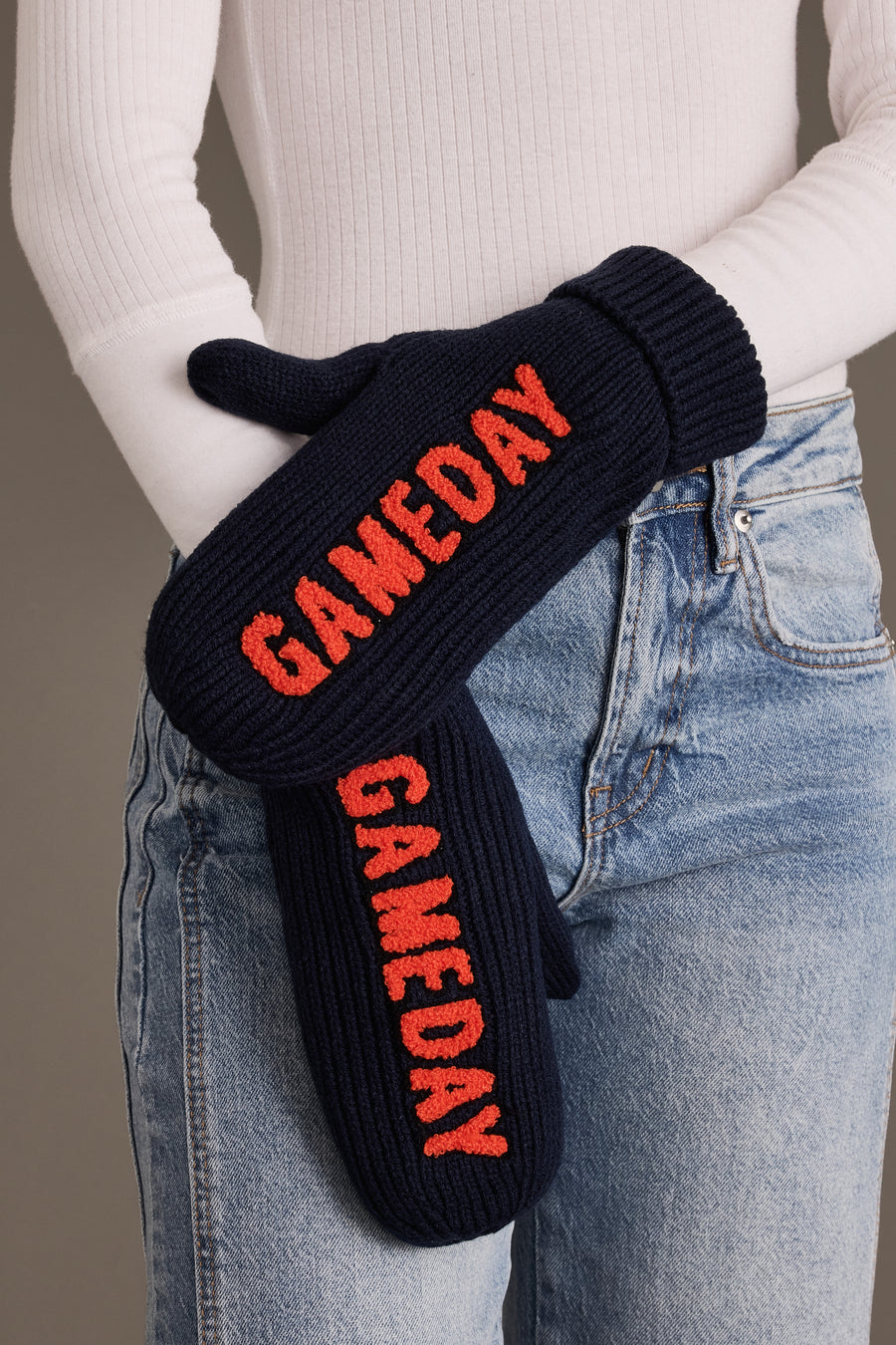 A person in blue jeans and a white long-sleeve top is holding Panache Apparel Collection’s Navy & Orange Game Day Mittens, which feature “GAMEDAY” knitted in red on the fleece-lined navy knit.