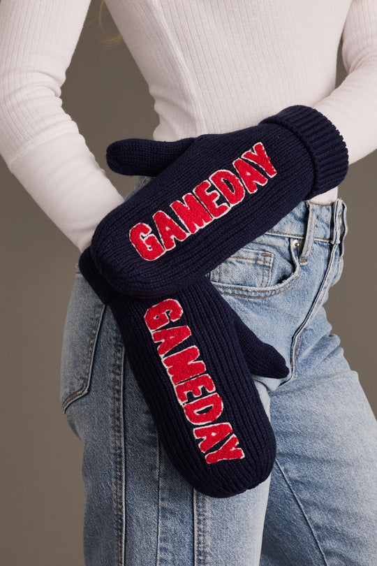 A person wearing blue jeans and a white ribbed turtleneck is shown with Panache Apparel Collection’s Navy & Red Game Day Mittens—fleece lined, navy blue knit mittens featuring bold red and white “GAMEDAY” lettering—resting on their hip.