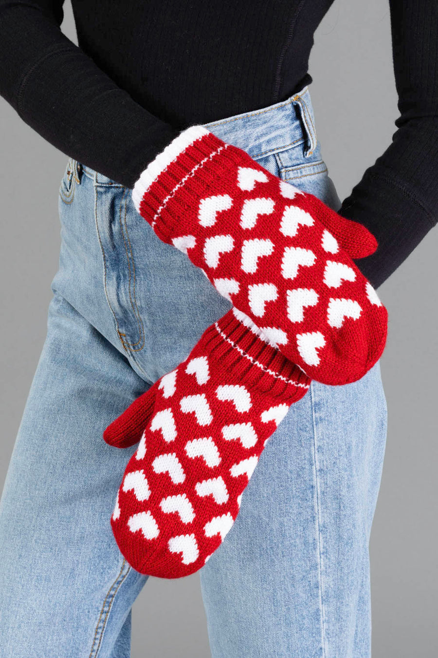 A person wearing blue jeans, a black sweater, and the Panache Apparel Collection Red & White Heart Pattern Mittens stands against a gray background.