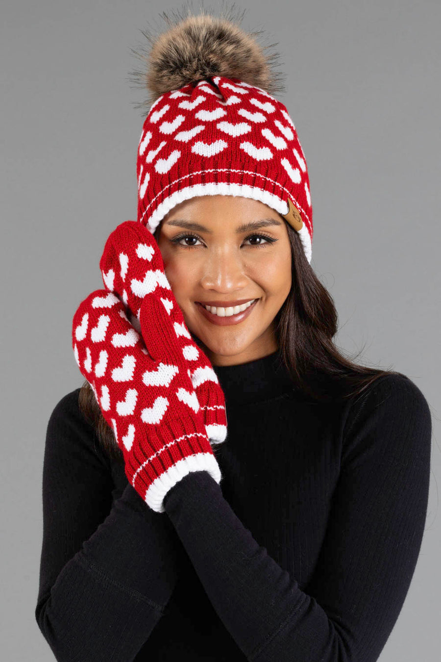 A smiling woman models the Panache Apparel Collection Red & White Heart Pattern Pom Hat, featuring a fur pom-pom, along with matching mittens and a black sweater, posed against a gray background.
