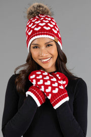 A woman smiles while wearing the Panache Apparel Collection’s Red & White Heart Pattern Pom Hat, featuring a fur pom-pom and matching mittens, paired with a black top and standing against a plain gray background.