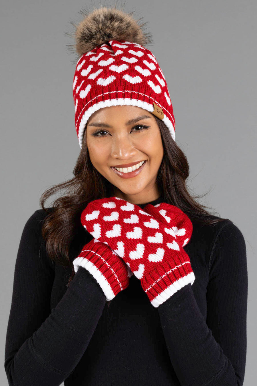A woman smiles while wearing the Panache Apparel Collection’s Red & White Heart Pattern Pom Hat, featuring a fur pom-pom and matching mittens, paired with a black top and standing against a plain gray background.