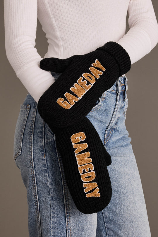 A person wearing a white ribbed long-sleeve top and light blue jeans holds the Panache Apparel Collection Black & Tan Game Day Mittens, featuring “GAMEDAY” embroidered in gold letters. These cozy mittens are fleece lined for extra warmth, making them perfect for chilly outdoor events.