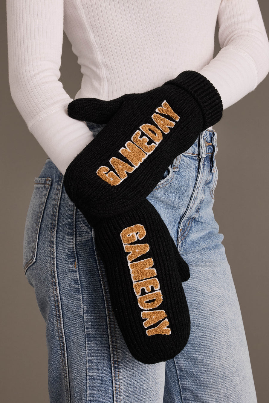 A person wearing a white ribbed long-sleeve top and light blue jeans holds the Panache Apparel Collection Black & Tan Game Day Mittens, featuring “GAMEDAY” embroidered in gold letters. These cozy mittens are fleece lined for extra warmth, making them perfect for chilly outdoor events.
