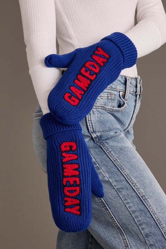 A person in a white sweater and blue jeans wears the Panache Apparel Collection Royal Blue & Red Game Day Mittens, fleece lined for extra warmth, with “GAMEDAY” in red letters on each mitten.