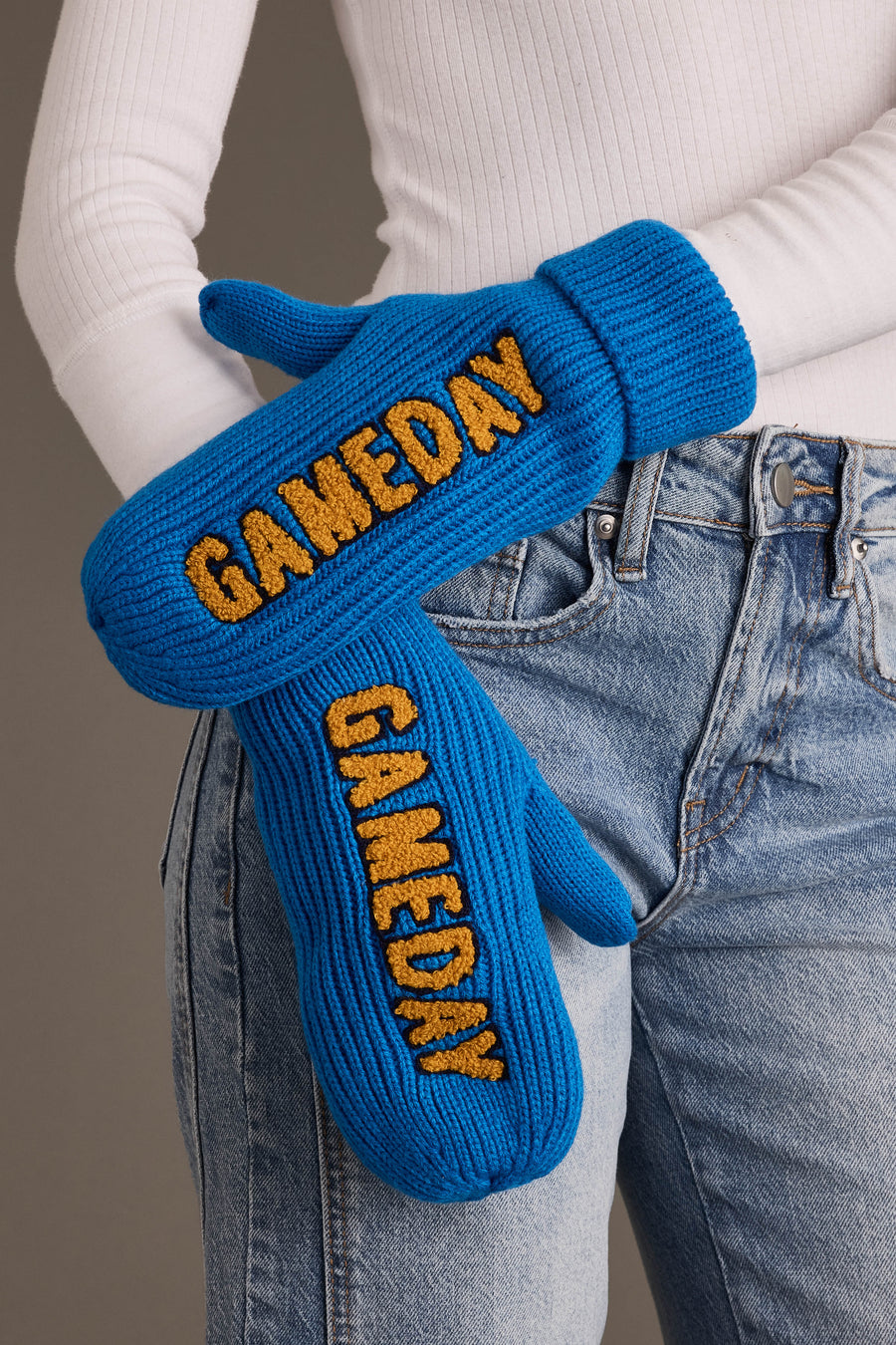 A person wearing Panache Apparel Collection’s Blue & Gold Game Day Mittens, featuring yellow “GAMEDAY” lettering, styled with light blue jeans and a white ribbed long-sleeve shirt.