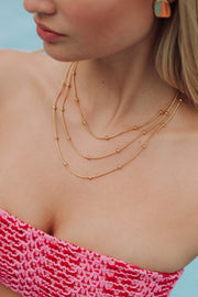 A woman wearing a pink crocheted strapless top, the Luxe Gold Layered Beaded Necklace by Panache Apparel Collection, and colorful round earrings, shown from shoulders to chin against a blue background.