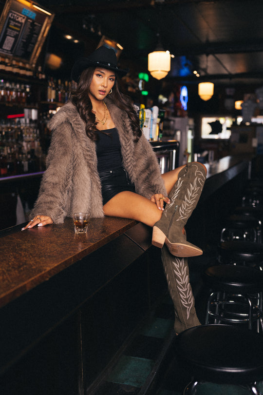 A woman wearing a fur coat, black cowboy hat, black shorts, and Panache Apparel Collection’s Olive Embroidered Western Boots sits on a bar counter with a drink in hand, set in a dimly lit bar.