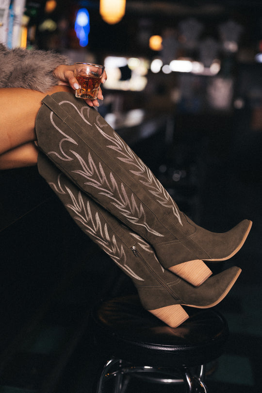 Wearing Panache Apparel Collection’s Olive Embroidered Western Boots, a person rests their legs on a bar counter while holding a glass of whiskey, set against the moody ambiance of a dimly lit bar.