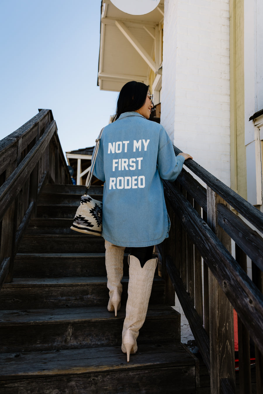 A woman wearing the Panache Apparel Collection’s Not My First Rodeo Button Up, white cowboy boots, and a patterned bag walks up a wooden outdoor staircase next to a white building.