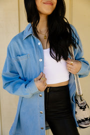 A woman styles the Not My First Rodeo Button Up by Panache Apparel Collection over a white ribbed crop top and black high-waisted pants, finished with gold rings, layered necklaces, and a patterned shoulder bag.