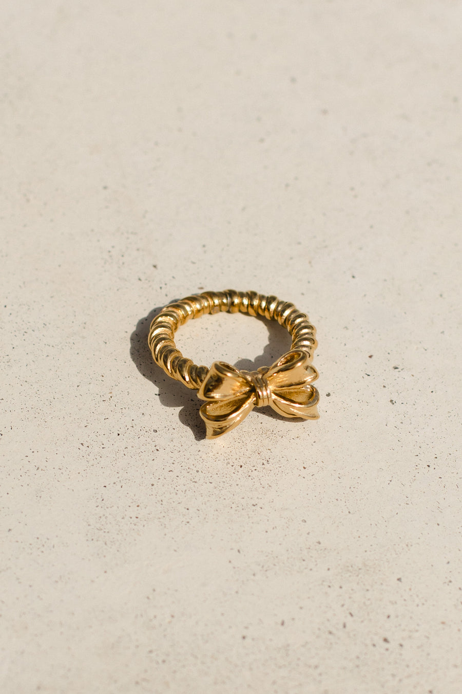 The Luxe Gold Textured Bow Ring by Panache Apparel Collection features an elegant 18K gold bow design with a ridged band, displayed on a light, speckled surface.