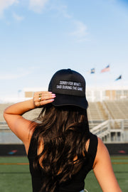 A woman with long brown hair stands before empty bleachers, wearing the Panache Apparel Collection's Sorry For What I Said During The Game Trucker Hat. She holds the brim of the adjustable snapback as two flags wave in the background beneath a bright blue sky.