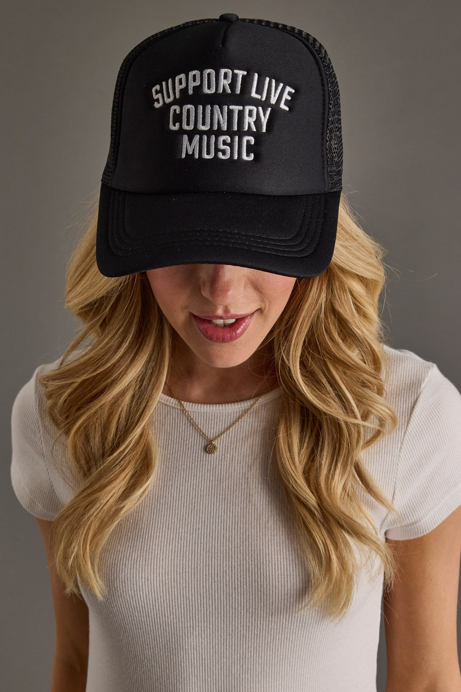 A person with long blonde hair wears a white ribbed shirt and the Panache Apparel Collection "Support Live Country Music" Trucker Hat, standing against a plain gray background.