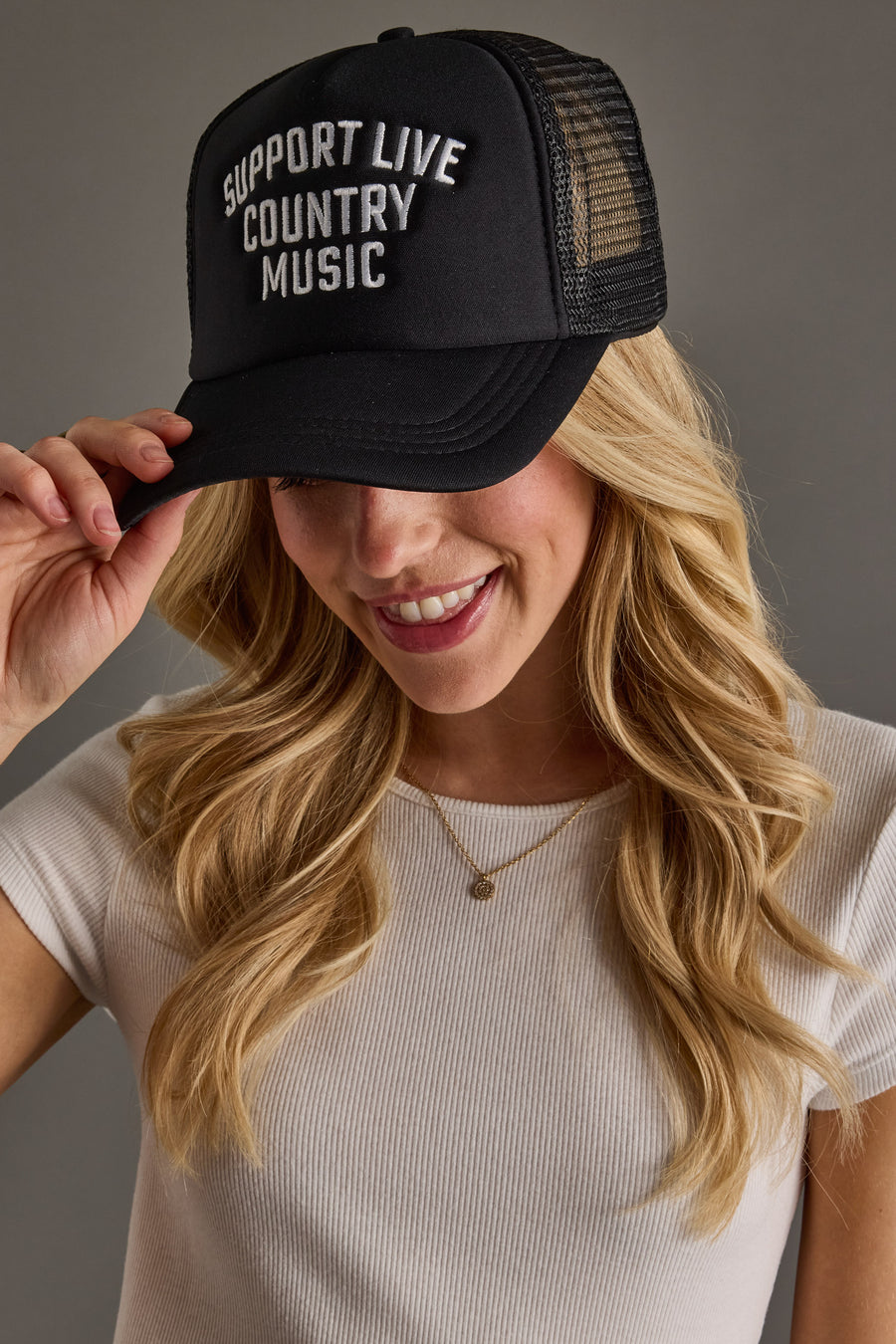 A smiling woman with long blonde hair wears a Panache Apparel Collection Support Live Country Music Trucker Hat and a ribbed white short-sleeve top with a gold necklace, holding the brim of the black snapback hat featuring white lettering.
