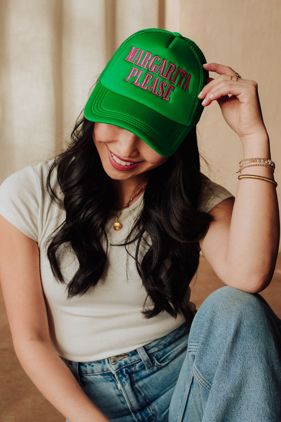 A woman with long dark hair and a white shirt smiles as she tilts the green Margarita Please Trucker Hat by Panache Apparel Collection, featuring red and pink lettering. She pairs it with jeans and gold bracelets.