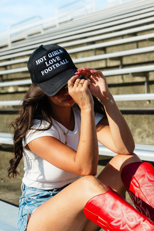 A woman in red boots and denim shorts sits on bleachers wearing the Panache Apparel Collection's Hot Girls Love Football Trucker Hat—a black cap with an adjustable snapback—and a white t-shirt, holding the brim of her hat and looking down.