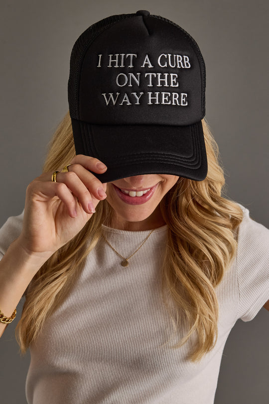 A smiling woman with long blonde hair wears the "I Hit A Curb On The Way Here" Trucker Hat from Panache Apparel Collection, holding its brim. She pairs it with a white ribbed shirt and gold jewelry.