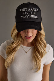 A woman with long blonde hair wears a Panache Apparel Collection I Hit A Curb On The Way Here Trucker Hat with white embroidered text, paired with a white ribbed shirt and gold necklace.