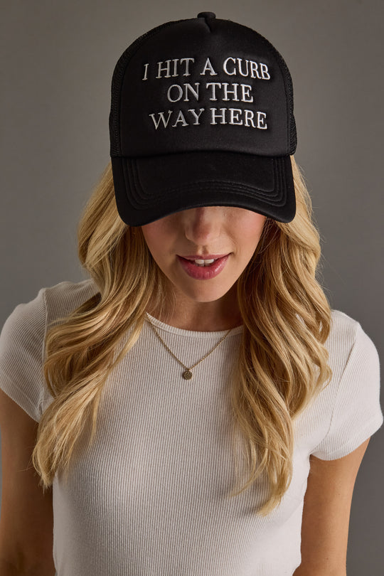 A woman with long blonde hair wears a Panache Apparel Collection I Hit A Curb On The Way Here Trucker Hat with white embroidered text, paired with a white ribbed shirt and gold necklace.