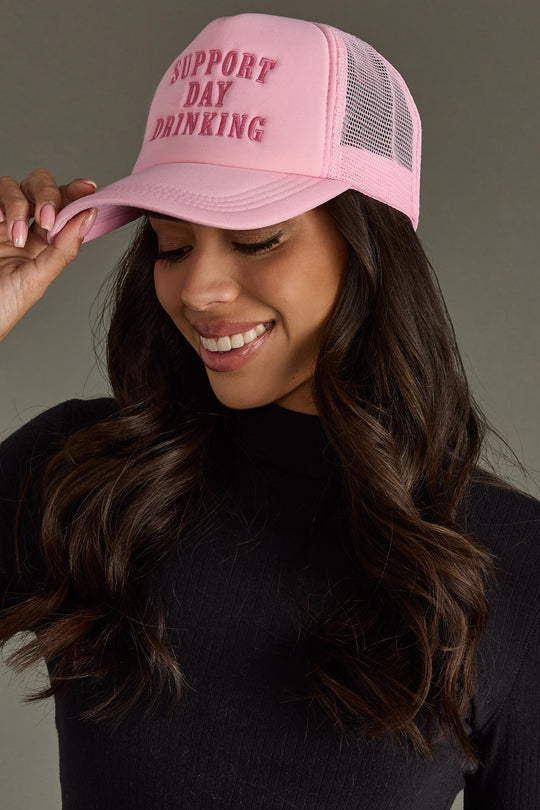 A smiling woman with long brown hair wears the Panache Apparel Collection's Support Day Drinking Trucker Hat in pink, paired with a black top as she touches the brim.