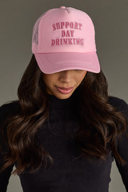 A woman with long dark hair wears the Panache Apparel Collection Support Day Drinking Trucker Hat and a black long-sleeve top, looking down with a neutral background.