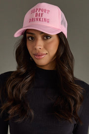 A woman with long, wavy brown hair wears the Panache Apparel Collection "Support Day Drinking" Trucker Hat and a black ribbed long-sleeve top, standing against a plain gray background.